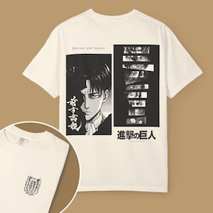 May include: An off-white short-sleeve t-shirt featuring a black and white graphic on the back with a character portrait, manga panels, and the text Dedicate your hearts and Attack on Titan. A small emblem is printed on the front chest area.