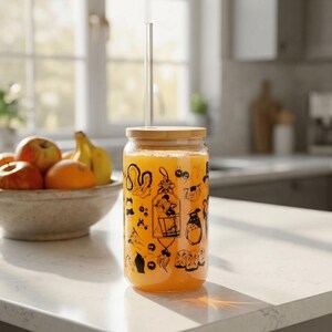 May include: A clear glass tumbler filled with orange liquid, featuring black line art illustrations of various animated characters. The glass has a bamboo lid and a straight clear glass straw inserted through the center of the lid.