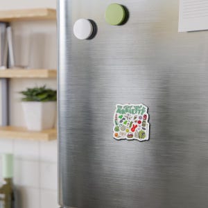 Op de afbeelding: Een rechthoekige magneet met een witte achtergrond, een groene rand en de tekst The Secret World of Arrietty. Het ontwerp bevat diverse kleine, kleurrijke illustraties van huishoudelijke voorwerpen, planten en een klein figuurtje in een groene jurk.