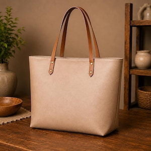 May include: A beige tote bag with two brown leather handles. The handles are attached to the top of the bag with gold-toned metal rivets. The bag has a structured rectangular shape with a smooth, matte finish and clean, minimalist stitching.