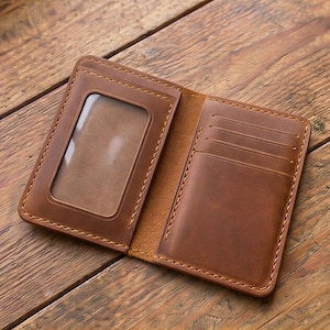 May include: Open, brown leather wallet with a clear ID window and multiple card slots. The wallet is stitched with a contrasting thread and lies open on a wooden surface, showcasing its interior design and craftsmanship.