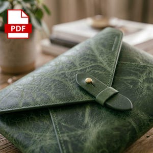 May include: A dark green leather envelope-style folder with a textured, distressed finish. The flap is secured by a matching green leather strap that loops through a small, centred brass-coloured metal stud button closure.