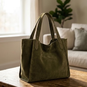 Puede incluir: Bolso tote de ante verde oliva con asas trenzadas. El bolso tiene forma rectangular con una abertura ancha y costuras visibles en los bordes. El bolso está sobre una mesa de madera.