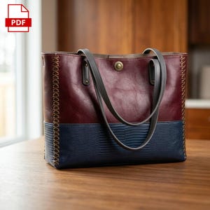 Peut inclure: Un sac fourre-tout en cuir bicolore. La partie supérieure est bordeaux, le bas est bleu marine avec un motif texturé. Le sac a des poignées grises et une fermeture à bouton. Les côtés sont cousus avec un fil décoratif. Le sac est posé sur une surface en bois.