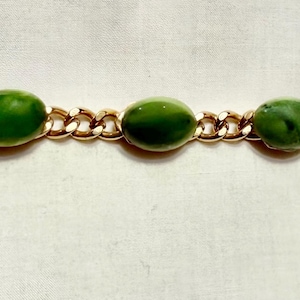 May include: A gold-toned bracelet featuring five oval, green gemstones. The gemstones are linked by a gold chain. The bracelet is displayed on a white surface. The clasp is visible at the end of the bracelet.