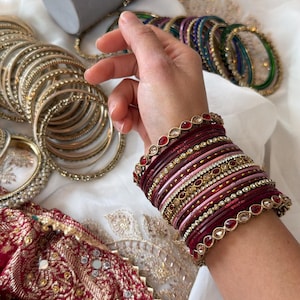May include: A stack of bangles in shades of deep red, pink, and gold, featuring metallic finishes, thin bands, and decorative edges with inset red stones. The set includes multiple textured and smooth rings worn together on the wrist.