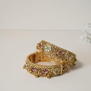 May include: Two gold-colored bangles with intricate floral and scrollwork designs. The bangles are adorned with small, colorful gemstones in shades of purple, green, and white. The jewelry is displayed on a white surface.