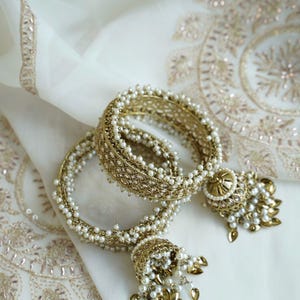 May include: Two gold-toned metal bangles featuring intricate filigree patterns, embellished with small white faux pearls along the edges. Each bangle includes a dangling bell-shaped charm adorned with matching pearls and small gold-toned leaf accents.