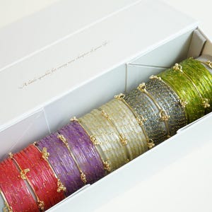 Könnte beinhalten: Eine weiße Schachtel mit einem Set bunter Armreifen. Die Armreifen sind in den Farbtönen Rot, Lila, Gold, Grau und Grün gehalten. Jeder Armreif hat goldene Akzente und kleine dekorative Elemente. Auf der Schachtel steht "A little sparkle for every new chapter of your life."