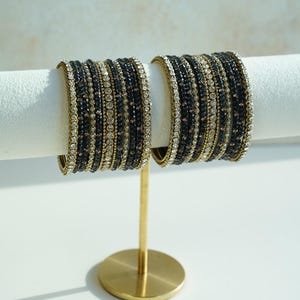 May include: Two sets of stacked bangles featuring alternating rows of dark black beads and clear sparkling rhinestones set in a gold-toned metal frame. The bangles are displayed on a gold-colored metal stand with a circular base and a horizontal bar.