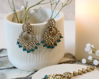 May include: A pair of gold-toned chandelier earrings and a matching maang tikka headpiece, both featuring intricate metalwork, clear white rhinestones, and small dangling teal-colored beads. The jewelry set displays a circular design.