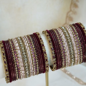 Peut inclure: Un ensemble de deux piles de bracelets joncs composé de bandes en velours bordeaux profond, en satin rose clair et en métal doré. L'ensemble comprend des joncs dorés ornés de petits cristaux transparents et de pierres ovales bordeaux.