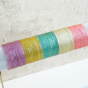 May include: A collection of colorful bangles in a rainbow of hues, including purple, yellow, teal, gold, and pink. The bangles are displayed on a white cylindrical stand against a neutral background. These beaded bracelets are a vibrant accessory.