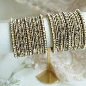 May include: A set of two stacks of gold-toned bangles featuring alternating rows of clear rhinestones and small white faux pearls. The bangles are arranged on a cylindrical display stand, showcasing a repeating pattern of metallic beads and stones.