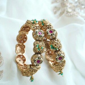 Peut inclure: Une paire de bracelets rigides dorés à charnières avec un travail de filigrane complexe. Chaque bracelet est orné de motifs floraux circulaires avec une pierre magenta centrale, entourée de petites perles blanches et de perles vertes, avec des pampilles.