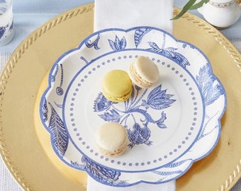 May include: A white paper plate with a blue floral pattern and scalloped edges. The plate is on top of a gold charger plate. There are three macarons on the plate, two are yellow and one is white.