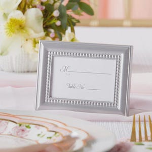 May include: A silver-colored picture frame with a beaded border, holding a white card with the letter "M" and the words "Table No." printed on it. The frame is set on a white tablecloth, with a floral arrangement in the background.