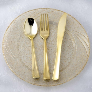 Pu&ograve; includere: Un set di cucchiaio, forchetta e coltello color oro su un piatto trasparente con macchie dorate e un bordo dorato. Le posate sono disposte sul piatto. Il piatto ha una superficie strutturata.