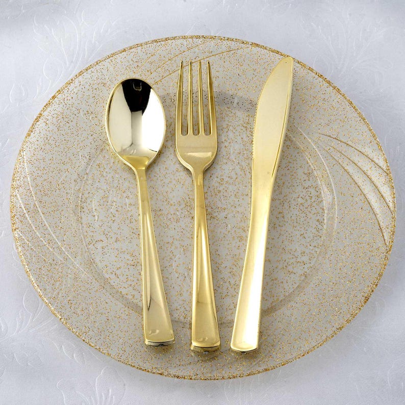Pu&ograve; includere: Un set di cucchiaio, forchetta e coltello color oro su un piatto trasparente con macchie dorate e un bordo dorato. Le posate sono disposte sul piatto. Il piatto ha una superficie strutturata.