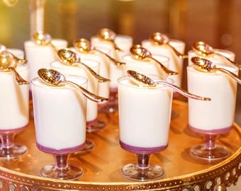 May include: A gold-colored tiered stand with a round top holds small clear glass cups filled with white dessert topped with a thin layer of purple. Each cup has a small gold spoon resting on the rim.