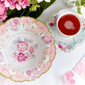 May include: A floral-themed tea party setting with a pink and white decorative plate, a matching teacup and saucer, and a cup of red liquid. The plate and saucer have a scalloped edge and gold trim. Pink flowers are in the background.