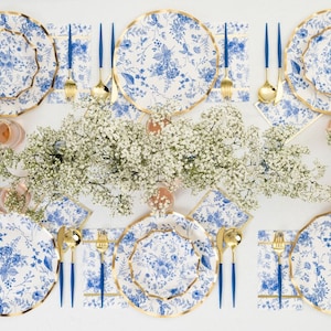 May include: A table setting with floral patterned plates, napkins, and gold-colored cutlery with blue handles. The plates have a gold trim. The table is decorated with white flowers. The plates and napkins have a blue floral design.