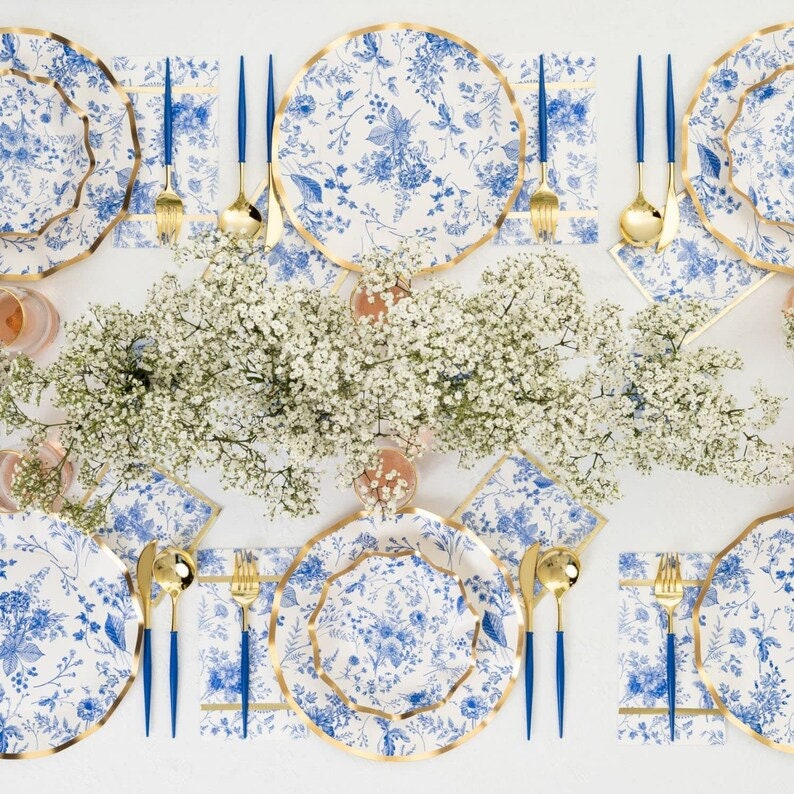 May include: A table setting with floral patterned plates, napkins, and gold-colored cutlery with blue handles. The plates have a gold trim. The table is decorated with white flowers. The plates and napkins have a blue floral design.