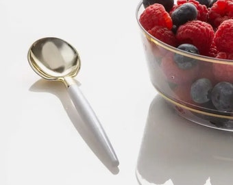 May include: A white and gold spoon with a gold bowl and a white handle. The spoon is lying on a white surface. A glass bowl filled with red raspberries and blueberries is in the background.