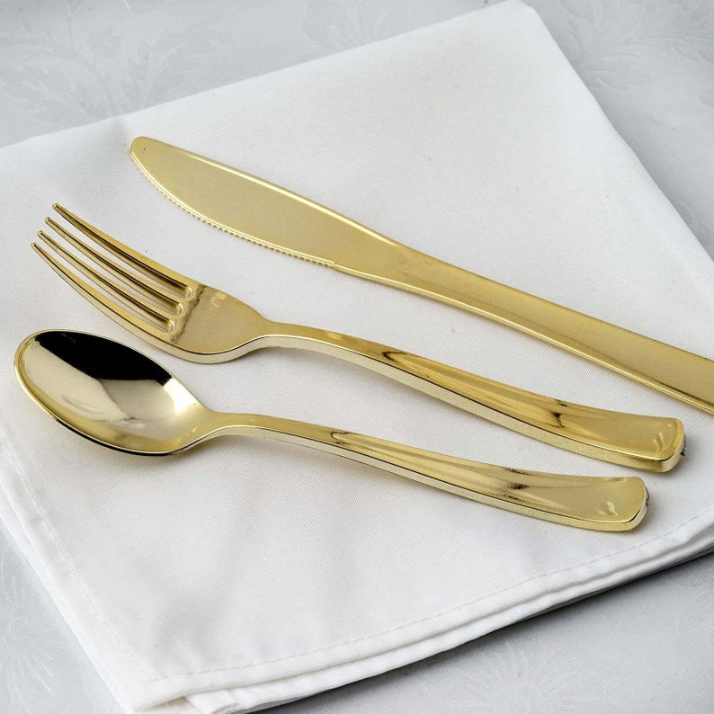 Pu&ograve; includere: Un set di posate color oro, tra cui forchetta, coltello e cucchiaio, &egrave; disposto su un panno bianco. Il coltello ha un bordo seghettato. Le posate hanno una superficie lucida e riflettente.