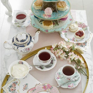 May include: A three-tiered cake stand with floral and bird designs, filled with scones and cupcakes. A teapot, teacups, and saucers are arranged on a gold tray with floral napkins and a plate of scones with cream and jam.