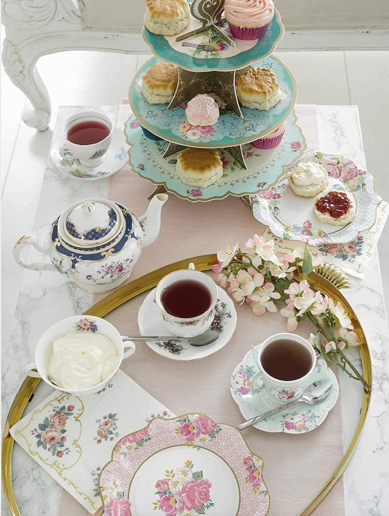 May include: A three-tiered cake stand with floral and bird designs, filled with scones and cupcakes. A teapot, teacups, and saucers are arranged on a gold tray with floral napkins and a plate of scones with cream and jam.