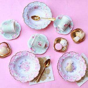May include: A collection of pastel-colored tea party supplies, including plates, cups, saucers, and spoons. The plates and cups feature floral designs with pink and blue accents. Gold-colored spoons and small serving dishes are also present, all arranged on a pink surface.