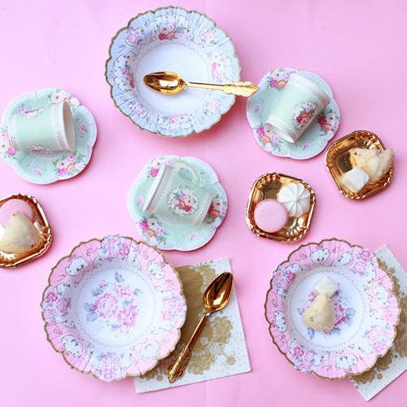 May include: A collection of pastel-colored tea party supplies, including plates, cups, saucers, and spoons. The plates and cups feature floral designs with pink and blue accents. Gold-colored spoons and small serving dishes are also present, all arranged on a pink surface.