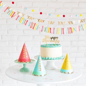 Peut inclure: Un g&acirc;teau blanc avec un gla&ccedil;age turquoise et un topper dor&eacute; "Joyeux anniversaire". Trois chapeaux de f&ecirc;te avec des lettres dor&eacute;es sont sur une table blanche avec un mur de briques blanches en arri&egrave;re-plan. Les chapeaux de f&ecirc;te sont rouge, bleu et jaune.