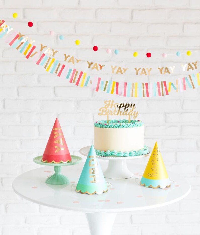 Peut inclure: Un g&acirc;teau blanc avec un gla&ccedil;age turquoise et un topper dor&eacute; "Joyeux anniversaire". Trois chapeaux de f&ecirc;te avec des lettres dor&eacute;es sont sur une table blanche avec un mur de briques blanches en arri&egrave;re-plan. Les chapeaux de f&ecirc;te sont rouge, bleu et jaune.
