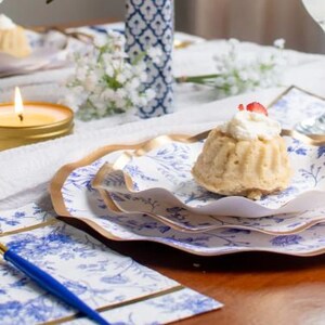 May include: A table setting with a dessert on a blue and white floral plate. A gold-colored fork with a blue handle rests on a matching napkin. A lit candle and a glass of water are also present.