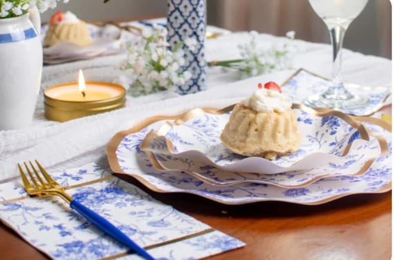 May include: A table setting with a dessert on a blue and white floral plate. A gold-colored fork with a blue handle rests on a matching napkin. A lit candle and a glass of water are also present.