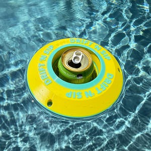 Könnte beinhalten: Ein gelber und türkisfarbener schwimmender Getränkehalter mit dem Text "DRIFT N SIP" schwimmt in einem Pool. Eine grüne Getränkedose befindet sich im Halter. Das Wasser ist klar blau.