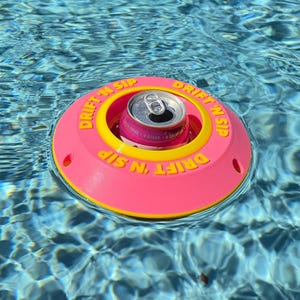Könnte beinhalten: Ein pinkfarbener und gelber schwimmender Getränkehalter mit den Worten "DRIFT 'N SIP" in Gelb. Eine Getränkedose befindet sich in der Mitte des Halters und schwimmt in einem Pool. Der Getränkehalter ist so konzipiert, dass Getränke schwimmen.