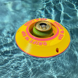 Puede incluir: Un portavasos flotante amarillo y rosa con el texto "DRIFT N SIP" en el borde. Una lata de bebida verde y dorada se encuentra en el centro del flotador, en una piscina.
