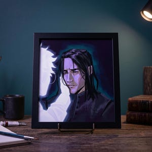 May include: A framed art print featuring a portrait of a person with long black hair and a somber expression. The artwork is set against a dark background with a white silhouette. The frame is black, and the print is displayed on a wooden surface.