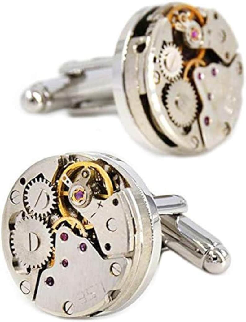 May include: A pair of silver cufflinks featuring the intricate internal mechanisms of a watch. The cufflinks showcase gears, jewels, and other components, offering a unique and mechanical aesthetic. The design includes silver-toned metal and small screws.