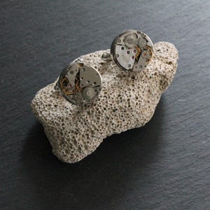 May include: A pair of silver cufflinks with exposed watch gears and intricate mechanical details. The round cufflinks are displayed on a textured, light-colored stone against a dark background. The gears are visible, showcasing the inner workings of a watch.