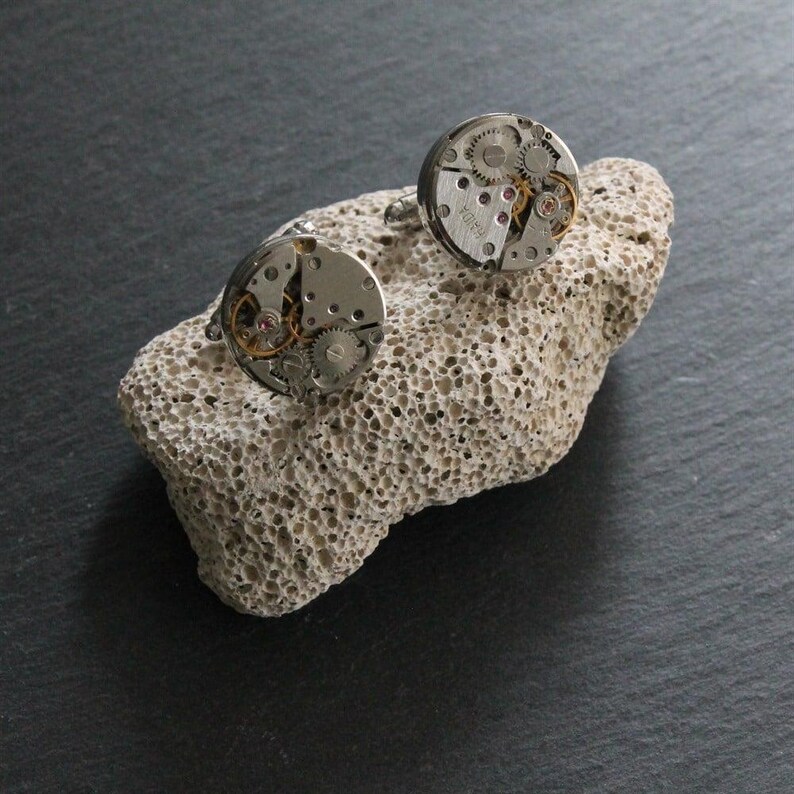May include: A pair of silver cufflinks with exposed watch gears and intricate mechanical details. The round cufflinks are displayed on a textured, light-colored stone against a dark background. The gears are visible, showcasing the inner workings of a watch.
