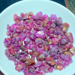 Puede incluir: Un cuenco de cerámica blanco lleno de una colección de gemas de rubí en bruto sin cortar. Los rubíes varían en tamaño y forma, mostrando tonos de rosa, rojo y púrpura. Algunas piedras tienen una superficie rugosa y texturizada.