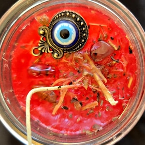 May include: A red candle in a glass jar, topped with an ornate, bronze-coloured charm featuring a blue eye design. The candle wax is embedded with herbs and small stones, and a white wick is visible.