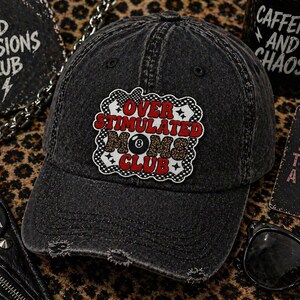 May include: A distressed black baseball cap with a patch that reads "OVER STIMULATED MOMS CLUB" in red, white, and black. The cap has a pre-worn look and is surrounded by other accessories.