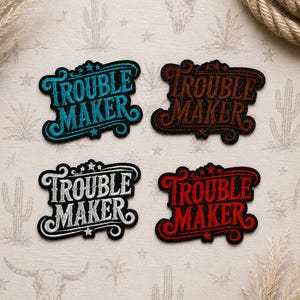May include: Four embroidered patches with the words "Trouble Maker" in a Western-style font. The patches are in teal, brown, white, and red, each with a decorative border and star accents. The patches are likely for clothing or accessories.