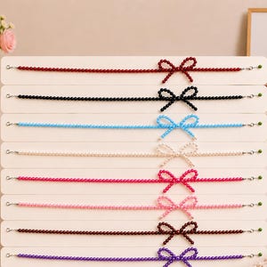 May include: Eight beaded necklaces displayed on a white board, each featuring a single row of small round beads in red, black, light blue, white, hot pink, light pink, dark brown, and purple, with each necklace tied into a bow shape.