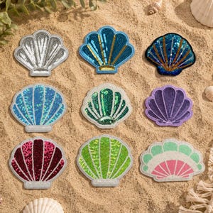 May include: Nine seashell-shaped patches in various colors, including silver, blue, green, purple, red, and pink. The patches are embellished with sequins and are arranged on a sandy surface with seashells and a starfish.